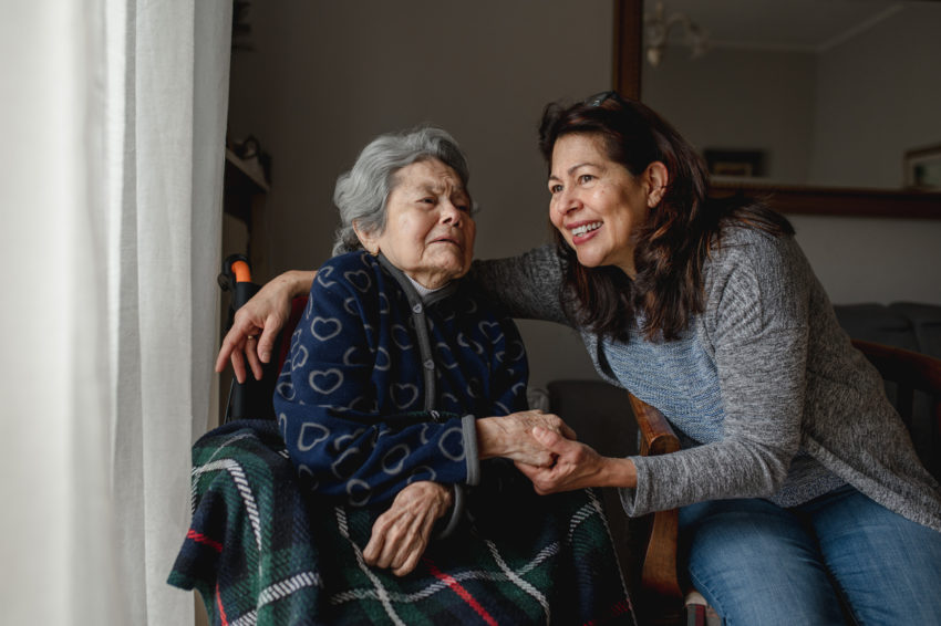 Old sick woman in wheelchair next to smiling daughter.